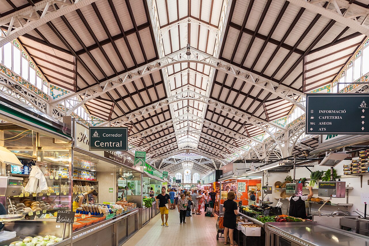 Shoppers and food stalls along the central aisle beneath Valencia Central Market's iron roof